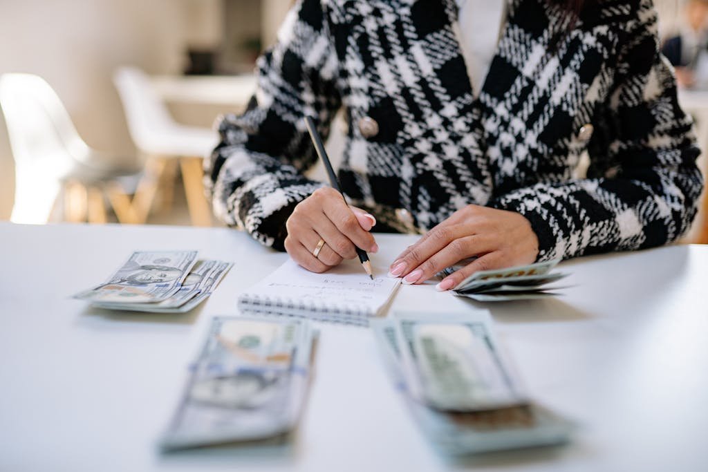 A woman in a plaid blazer writes in a notepad with cash spread on a white table, focusing on financial planning.