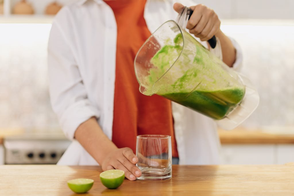 Close-up of a person pouring a vibrant green smoothie into a glass with limes on the wooden table.