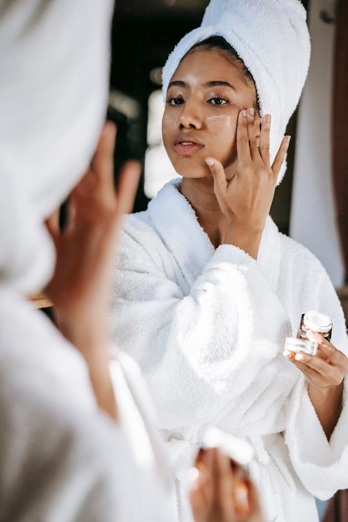 Young black female in white robe and towel on head applying moisturizing cream on face while standing in bathroom