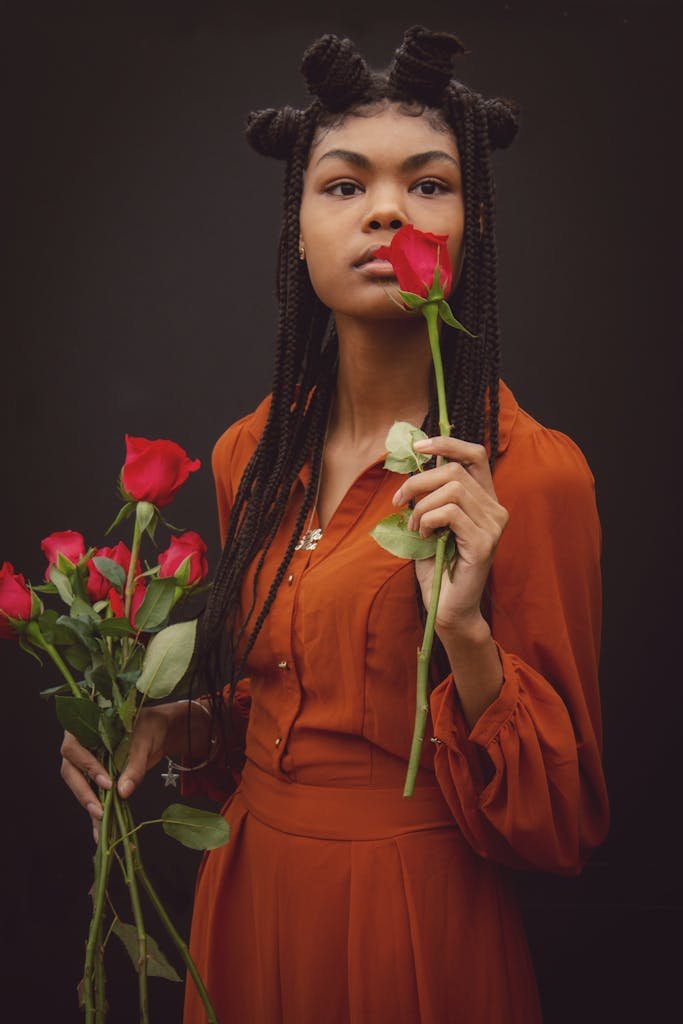 A stylish woman in an orange dress holds red roses against a black background.