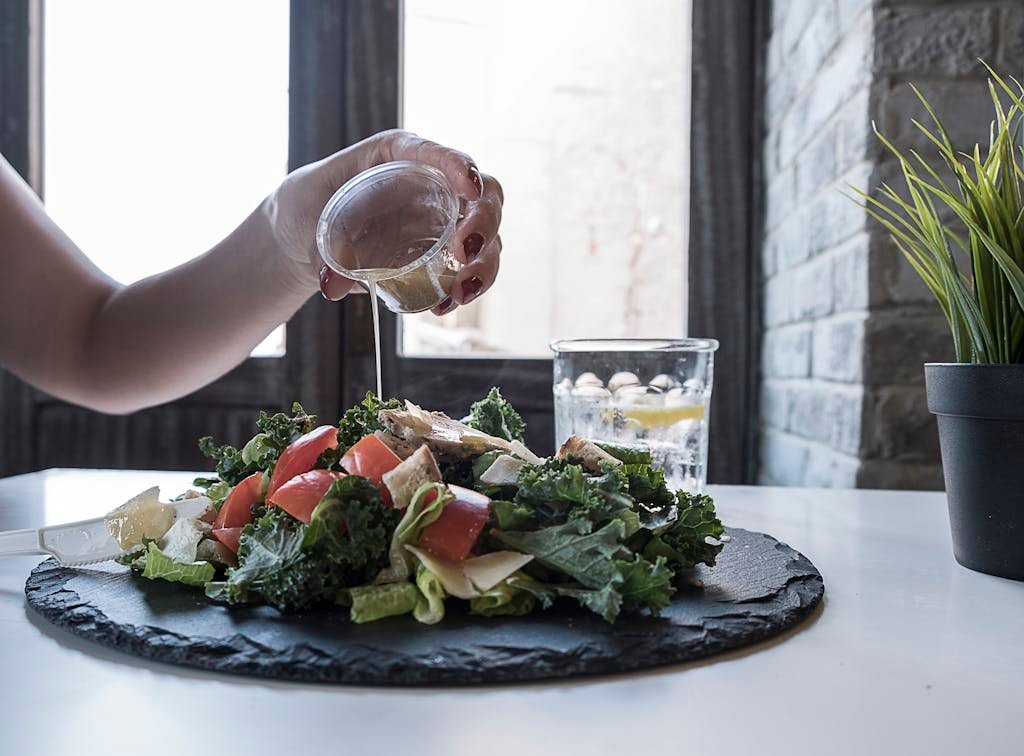 Hand pouring dressing over fresh salad with kale and tomatoes in a modern Dubai restaurant.