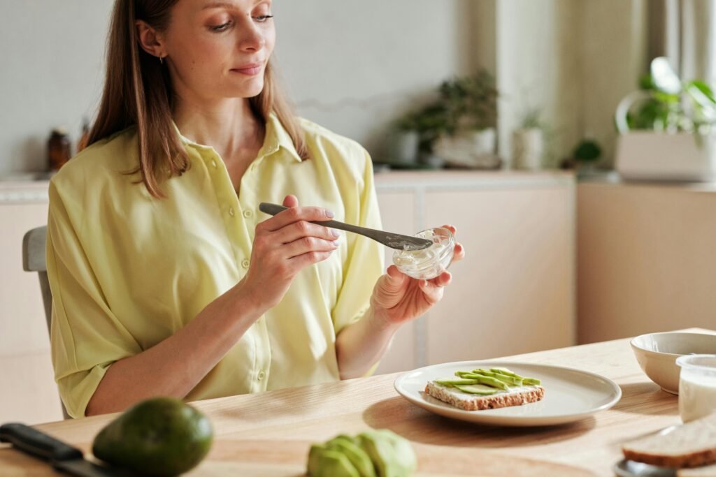 Young woman spreading cream cheese on toast with sliced avocado, creating a healthy breakfast.