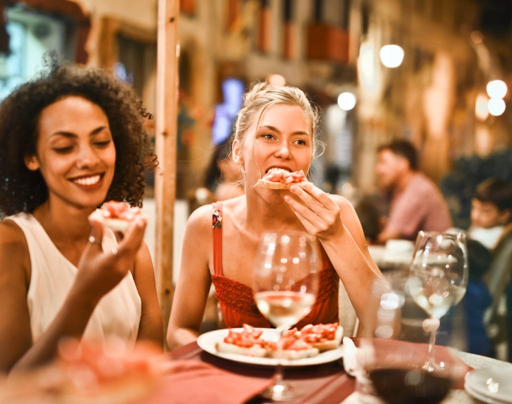 Two women enjoying bruschetta and wine at a vibrant outdoor restaurant.